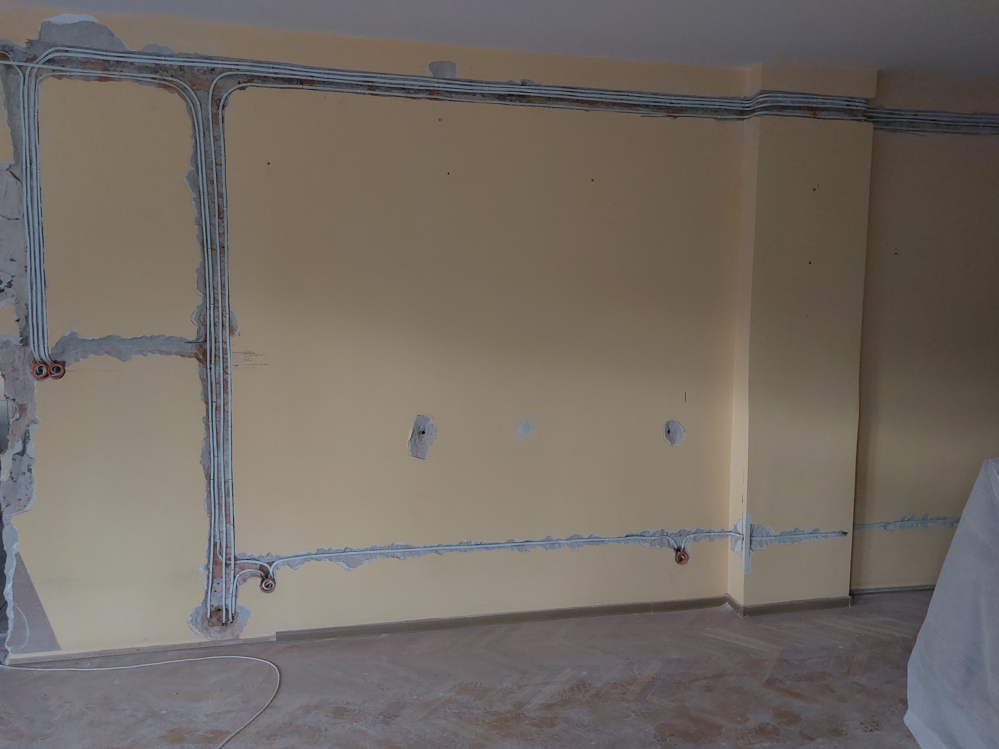 Wnętrze w trakcie remontu z widocznymi przewodami elektrycznymi w białych rurkach ułożonymi wzdłuż ścian, częściowo zamaskowanymi gipsem, z widocznymi puszkami instalacyjnymi.