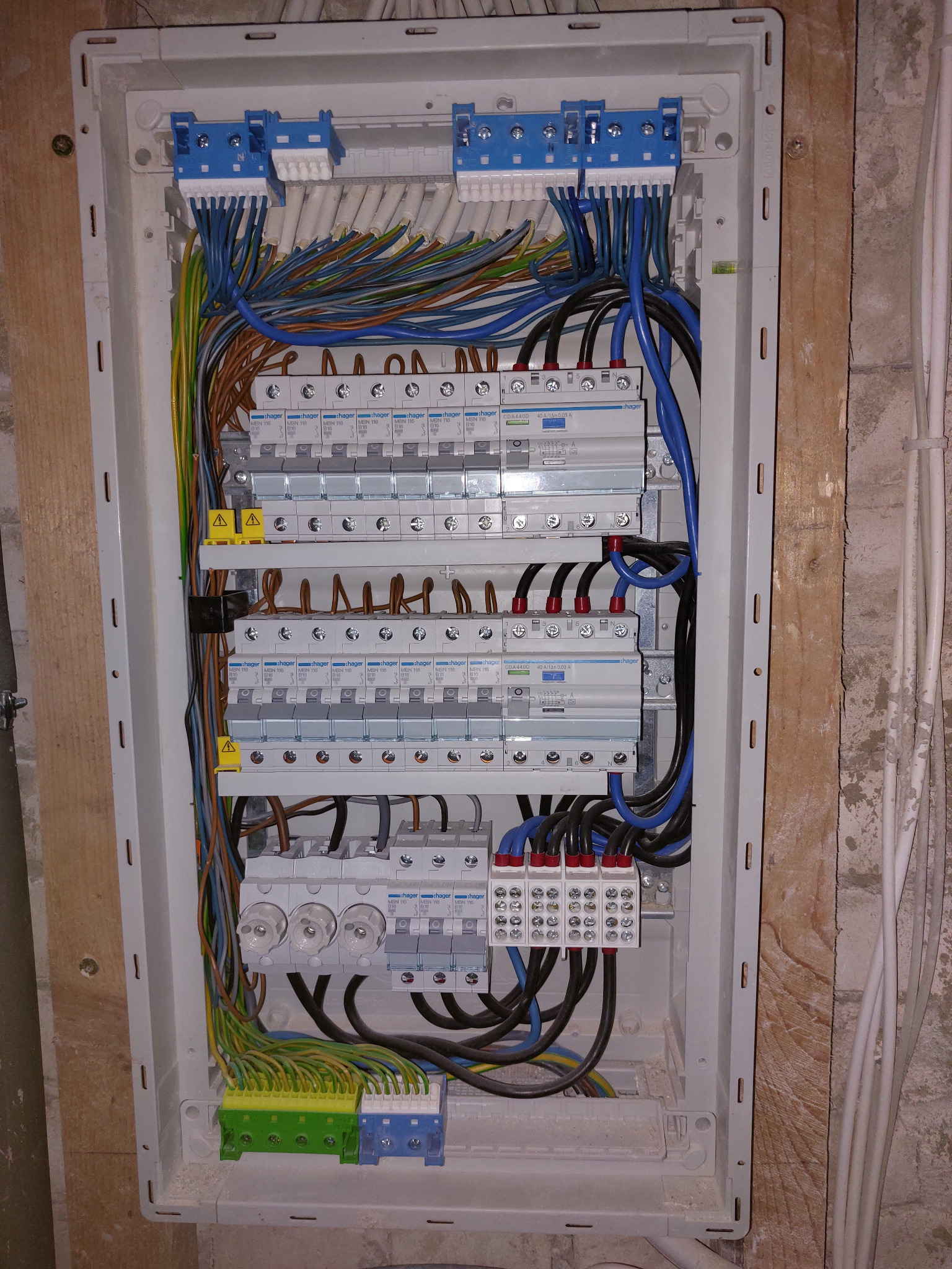 Otwarta skrzynka elektryczna z zainstalowanymi bezpiecznikami, przewodami o różnych kolorach i złączkami, zamontowana na ścianie z surowego drewna i betonu.