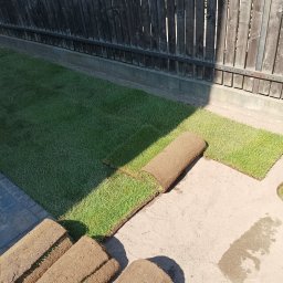 ROMANI GREEN GARDEN ROMAN ROHOVTSOV - Rozwinięta i nierozwinięta trawa w rolkach na betonowej powierzchni, widoczne ciemne drewniane ogrodzenie w tle.