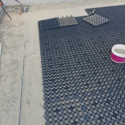 ROMANI GREEN GARDEN ROMAN ROHOVTSOV - Układanie plastikowej kratki trawnikowej na podsypce piaskowej, widoczny fragment kratki, wiadro z piaskiem i narzędzia.