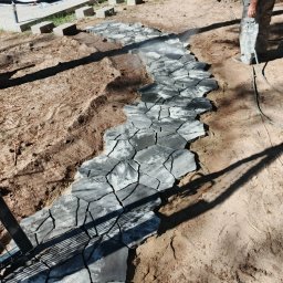 RGarden Green - Układanie nieregularnej ścieżki z szarych kamieni na piaszczystym podłożu, z widocznym fragmentem namiotu w tle i mężczyzną spryskującym kamienie wodą.