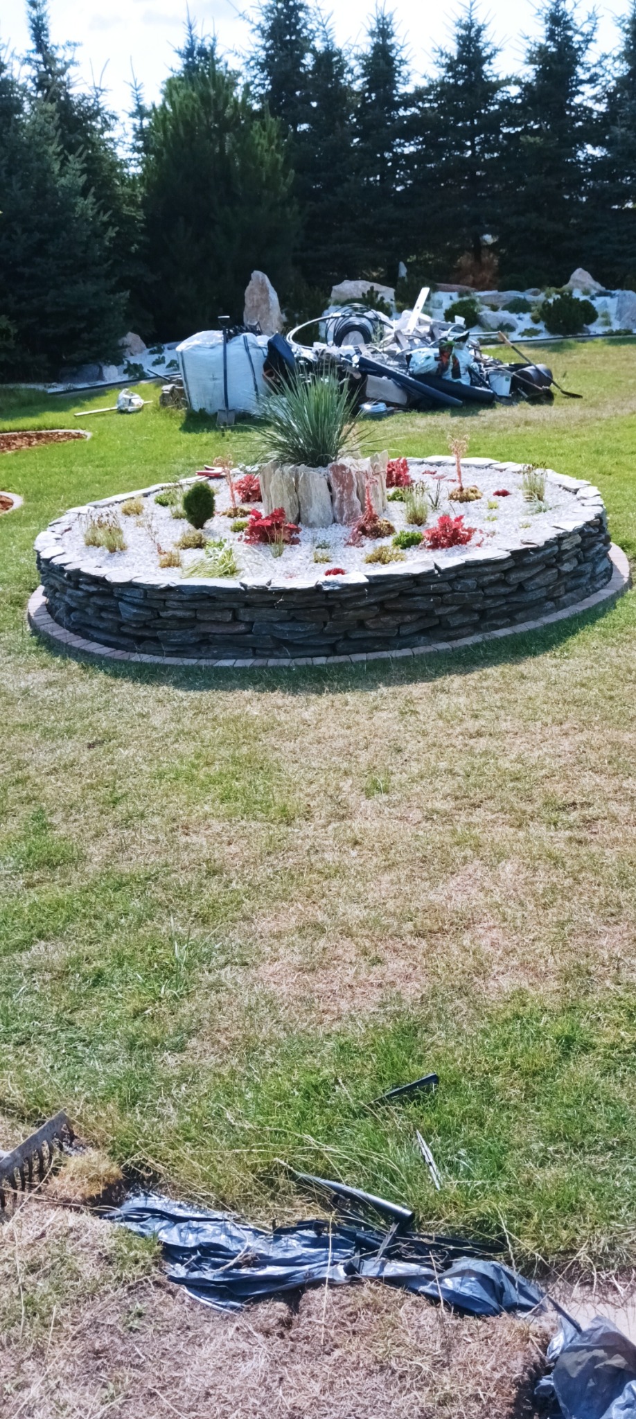 Okrągła rabata z kamienia i białego żwiru w Załuskach, z nasadzeniami roślin ozdobnych i centralną kompozycją z kamieni, otoczona trawnikiem. W tle widoczne drzewa iglaste i sprzęt ogrodniczy.