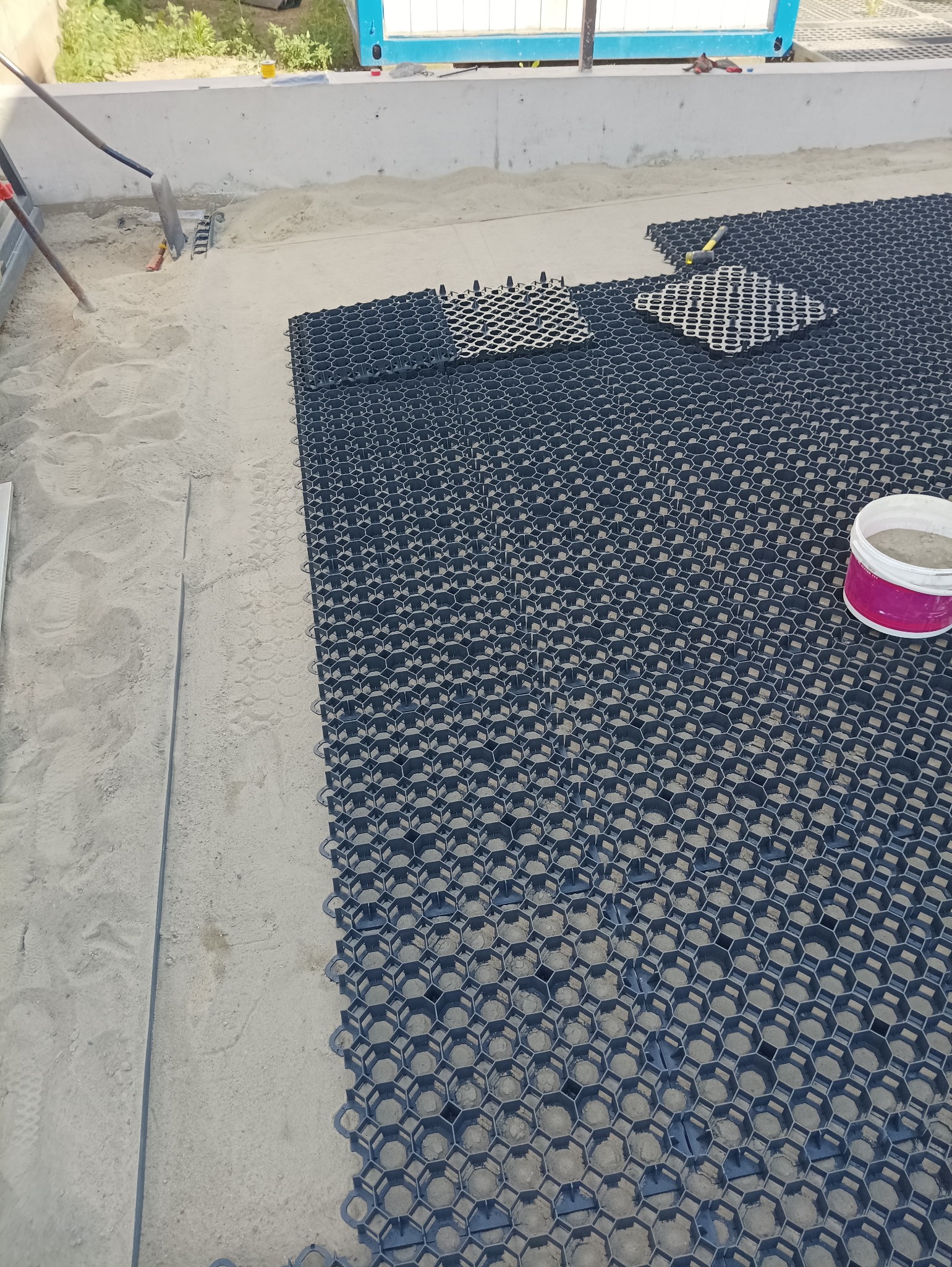 Układanie plastikowej kratki trawnikowej na podsypce piaskowej, widoczny fragment kratki, wiadro z piaskiem i narzędzia.