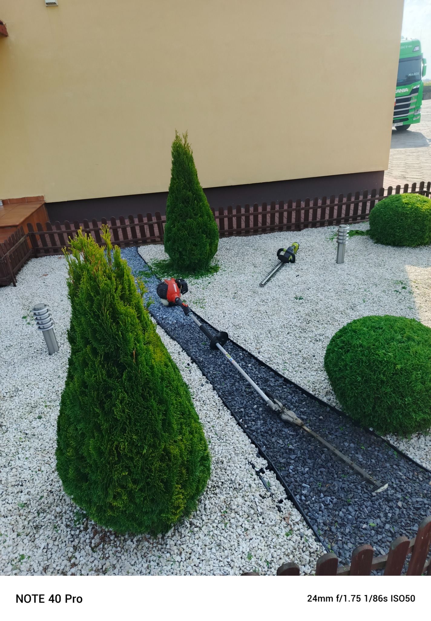 Widok z góry na ogród z ozdobnymi krzewami i ścieżką z kamyków, na której leżą narzędzia do formowania roślin, w tle ściana budynku i fragment ciężarówki.