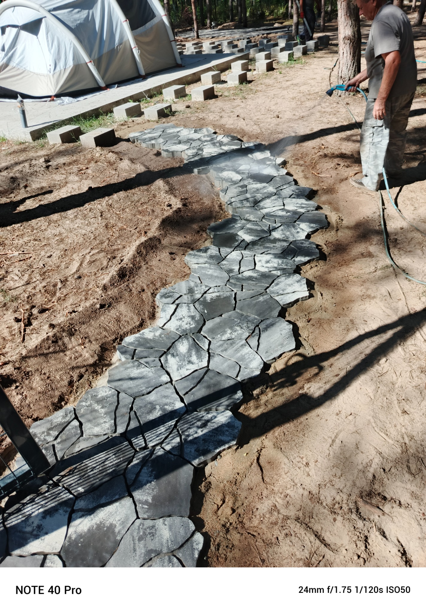 Układanie nieregularnej ścieżki z szarych kamieni na piaszczystym podłożu, z widocznym fragmentem namiotu w tle i mężczyzną spryskującym kamienie wodą.