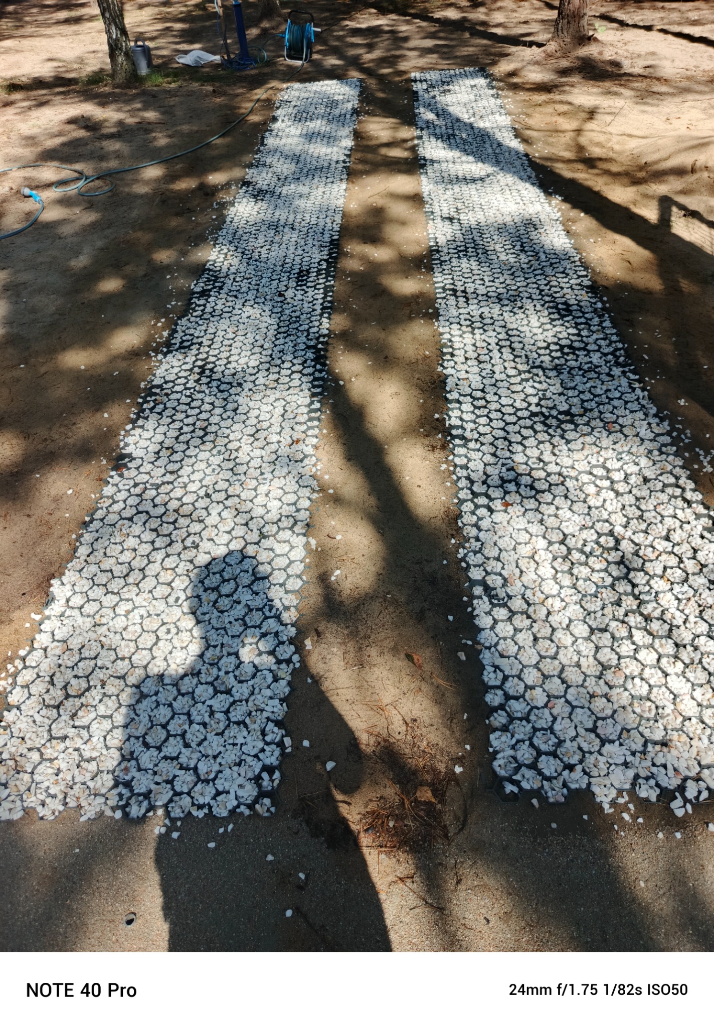 Dwie ścieżki z białego żwiru w czarnej siatce ułożone na piaszczystym podłożu w ogrodzie, z widocznymi cieniami drzew.