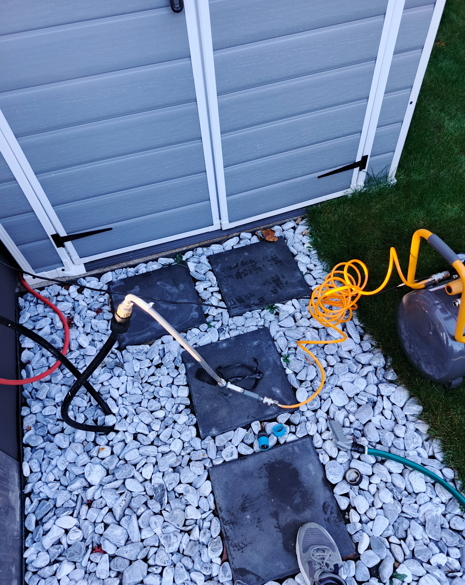 Szary domek narzędziowy otoczony białymi kamieniami i czarnymi płytami chodnikowymi, z widocznym kompresorem, wężem i elementami systemu nawadniania