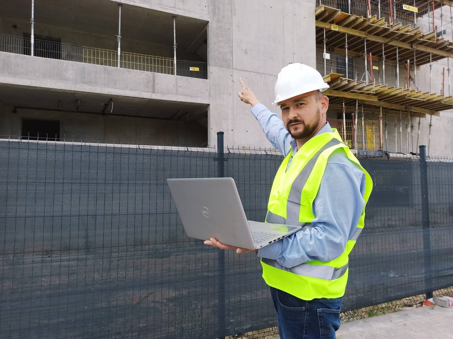 Mężczyzna w kasku i kamizelce odblaskowej, trzymający laptopa, wskazuje palcem na surowy budynek w tle.