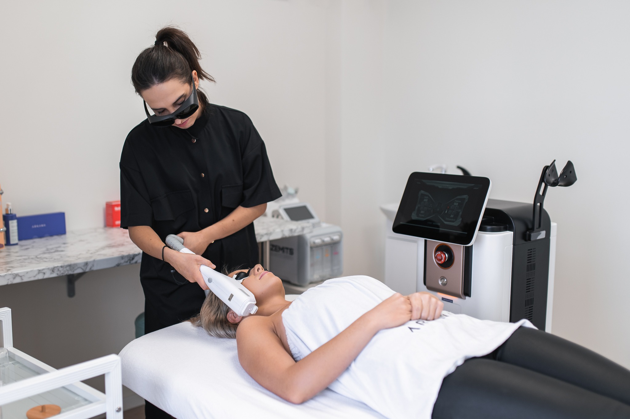 Kobieta w czarnym uniformie i okularach ochronnych wykonuje zabieg laserowy na twarzy klientki leżącej na białym łóżku kosmetycznym; w tle widoczny nowoczesny sprzęt kosmetyczny i marmurowy blat.