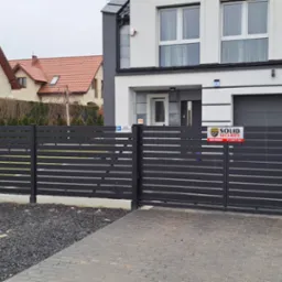 Nowoczesna, pozioma, metalowa brama wjazdowa i przęsła ogrodzeniowe w kolorze grafitowym, z widocznymi tabliczkami informacyjnymi i skrzynką pocztową, na tle dwóch domów.