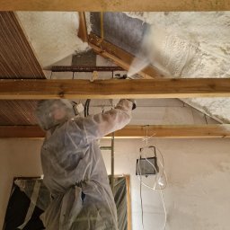 Eko Termika Kamil Jarosz - Pracownik w kombinezonie ochronnym aplikuje piankę izolacyjną na poddaszu, widoczne drewniane elementy konstrukcyjne dachu i fragmenty ocieplonej powierzchni.