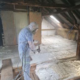 Eko Termika Kamil Jarosz - Pracownik w kombinezonie ochronnym aplikuje piankę PUR na poddaszu, widoczne drewniane elementy konstrukcji dachu i fragmenty ścian, jasne światło wpada przez okno dachowe.
