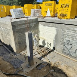 Eko Termika Kamil Jarosz - Fundament budynku w trakcie ocieplania pianką natryskową; widoczne żółte opakowania materiałów budowlanych Ytong, pracownik w kombinezonie ochronnym na drabinie, świeżo nałożona warstwa izolacji.