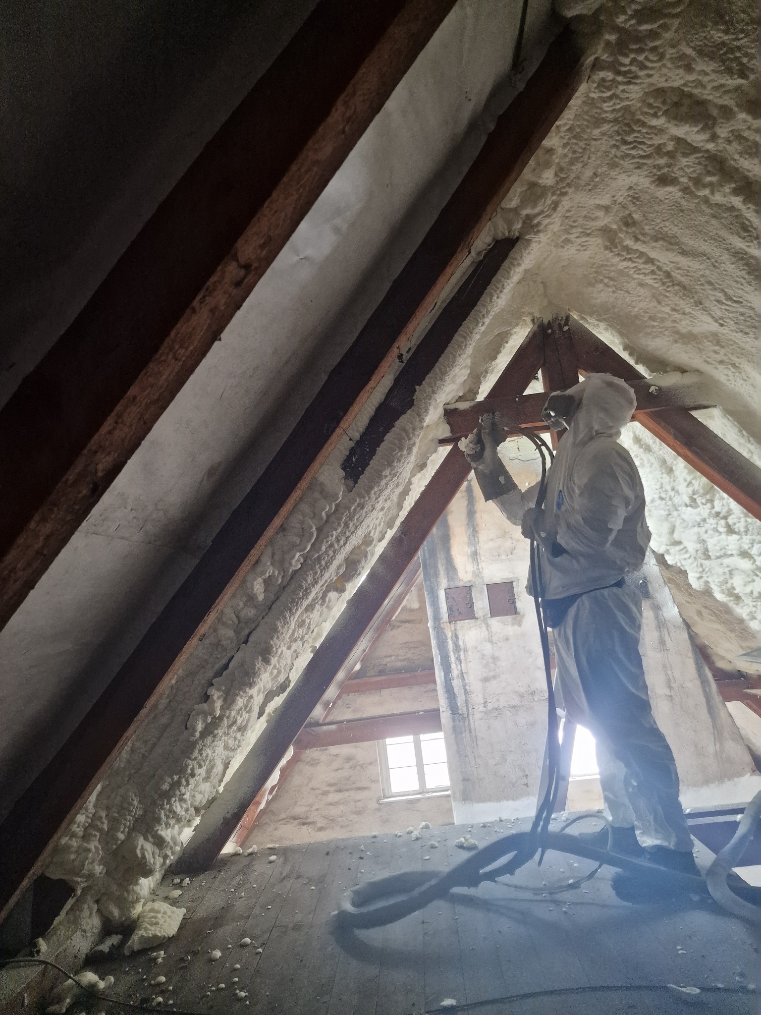 Pracownik w kombinezonie ochronnym natryskuje pianę izolacyjną na skośne poddasze, widoczne drewniane krokwie i fragment okna w tle.