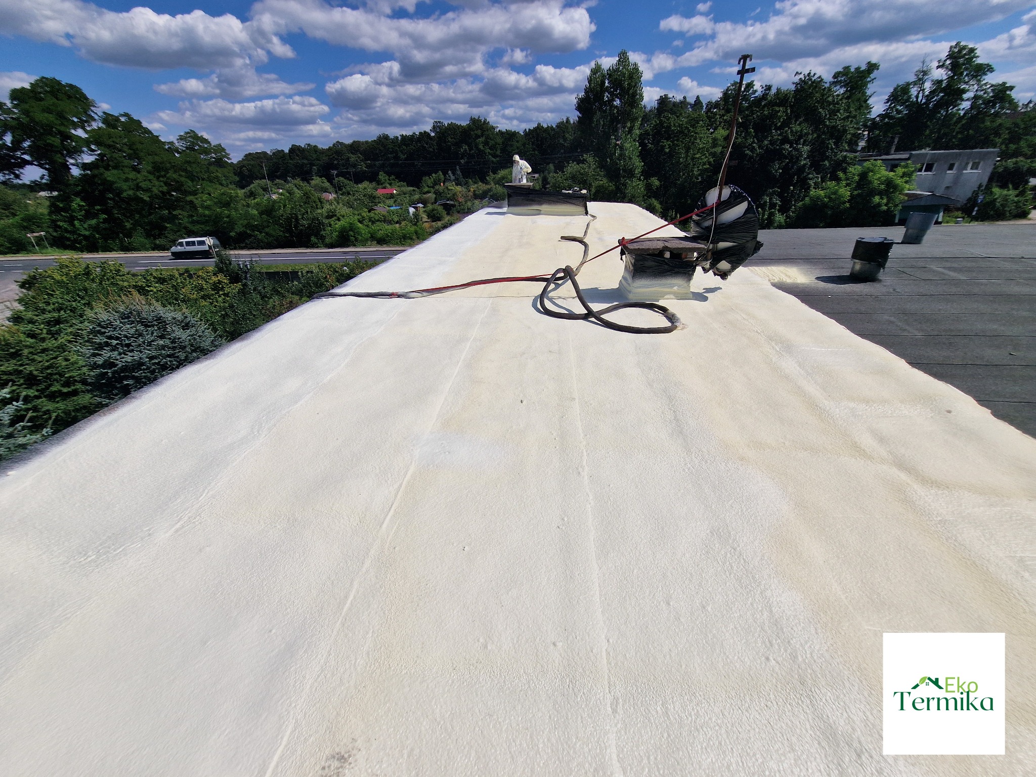 Biała pianka izolacyjna nanoszona na płaski dach budynku, widok z góry, w tle zieleń drzew i niebo z chmurami, widoczny pracownik w kombinezonie ochronnym i sprzęt do natrysku pianki.