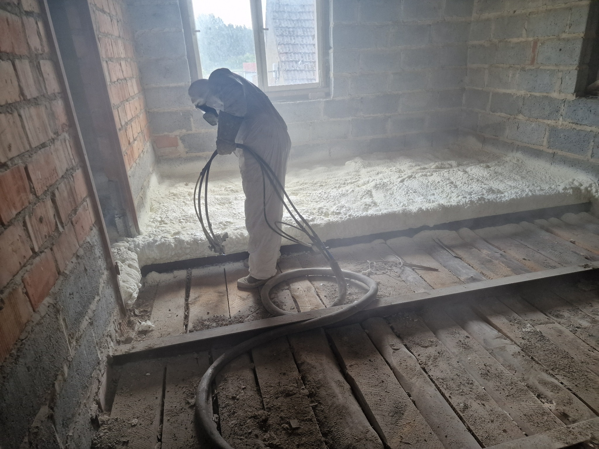 Pracownik w kombinezonie ochronnym aplikuje pianę izolacyjną na drewnianą podłogę poddasza w surowym stanie, widoczne ceglane ściany i okno w tle.