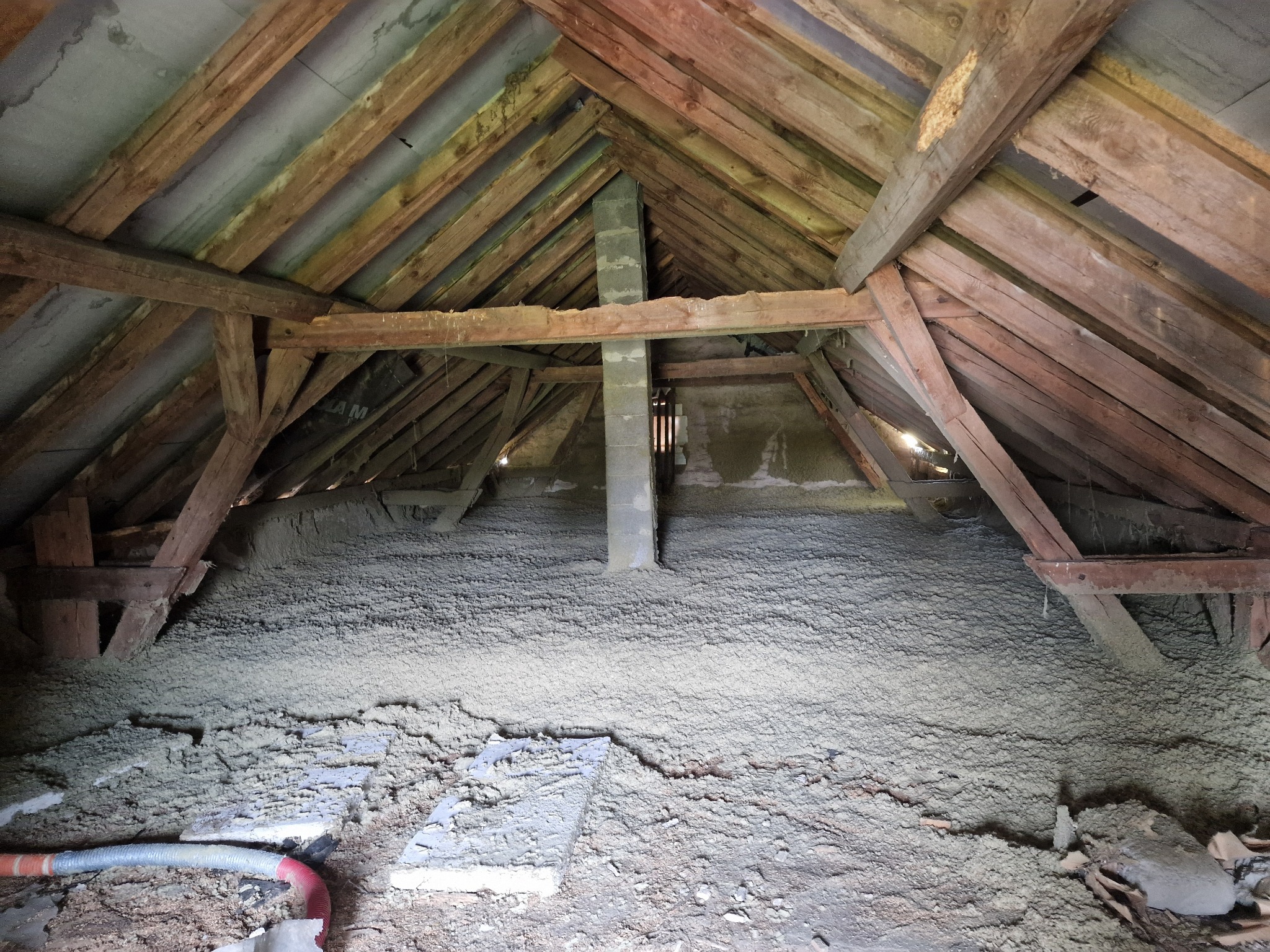 Poddasze z widoczną drewnianą konstrukcją dachu i warstwą izolacji z szarej pianki, z fragmentem przewodu do aplikacji izolacji w dolnym lewym rogu.