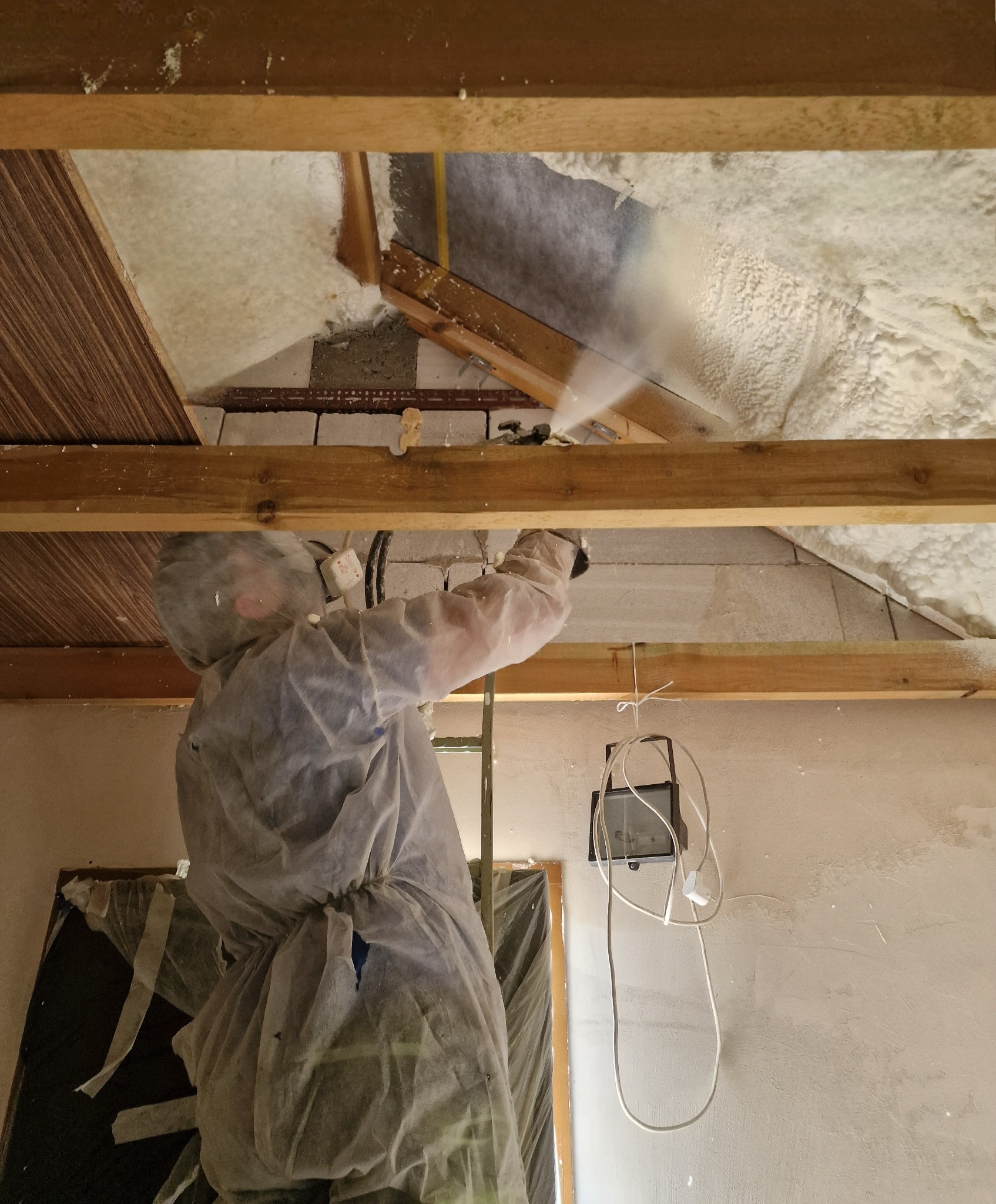Pracownik w kombinezonie ochronnym aplikuje piankę izolacyjną na poddaszu, widoczne drewniane elementy konstrukcyjne dachu i fragmenty ocieplonej powierzchni.