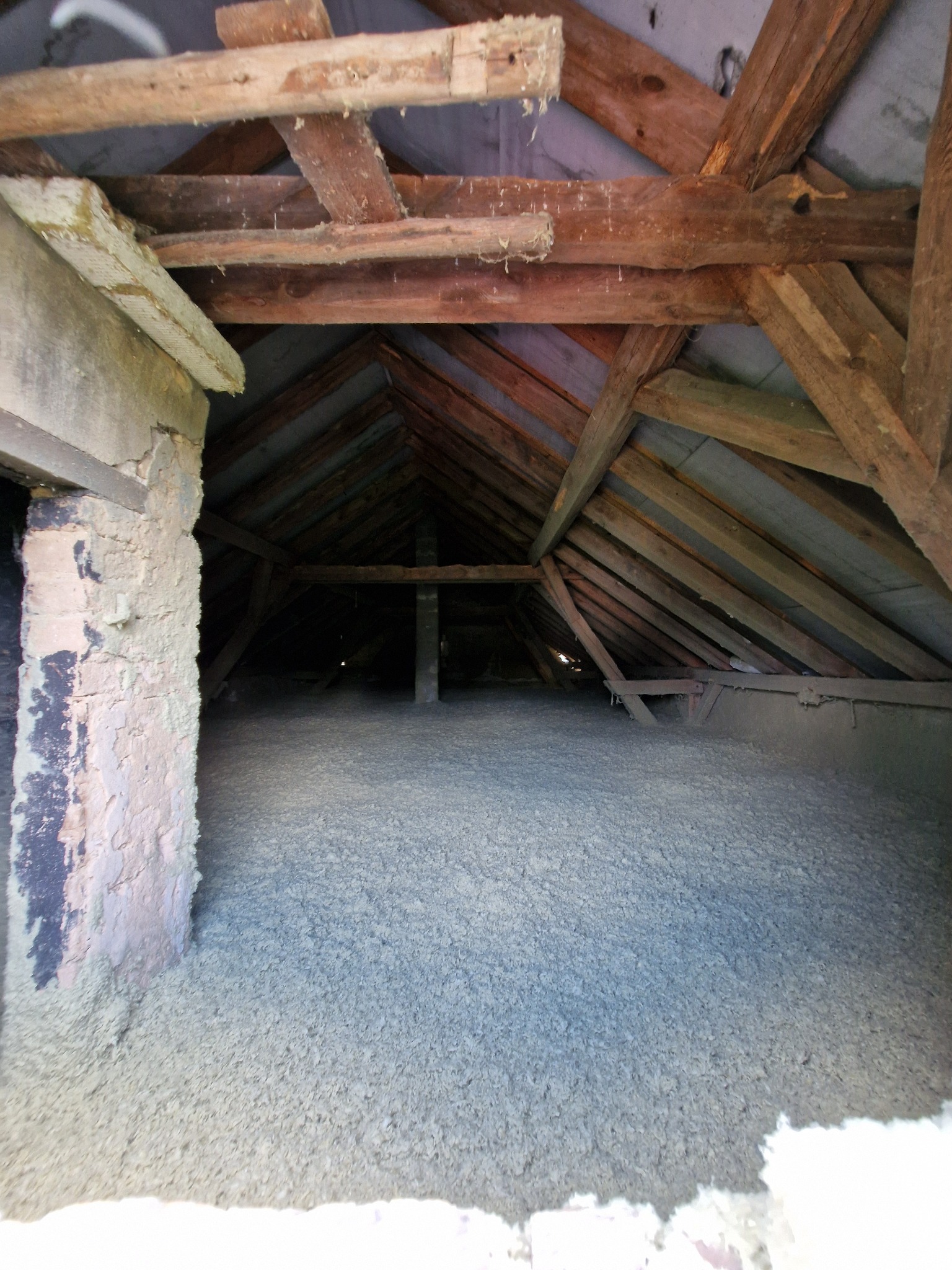 Poddasze z widoczną drewnianą konstrukcją dachu i warstwą szarej izolacji termicznej, prawdopodobnie wełny mineralnej, rozłożonej na podłodze.