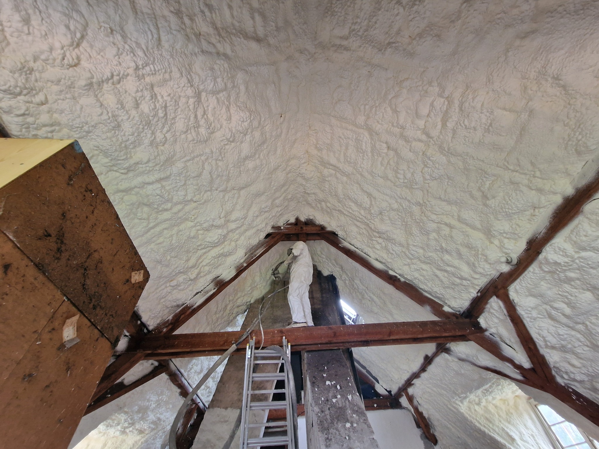 Pracownik w kombinezonie ochronnym natryskuje pianę izolacyjną na poddasze z drewnianą konstrukcją, widoczna drabina i elementy instalacji natryskowej.