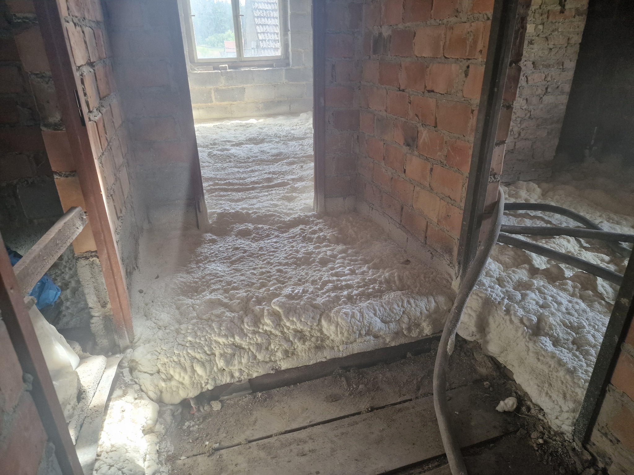 Poddasze w trakcie ocieplania: warstwa białej piany izolacyjnej pokrywa drewnianą podłogę w surowym wnętrzu z ceglanymi ścianami i oknem w tle.