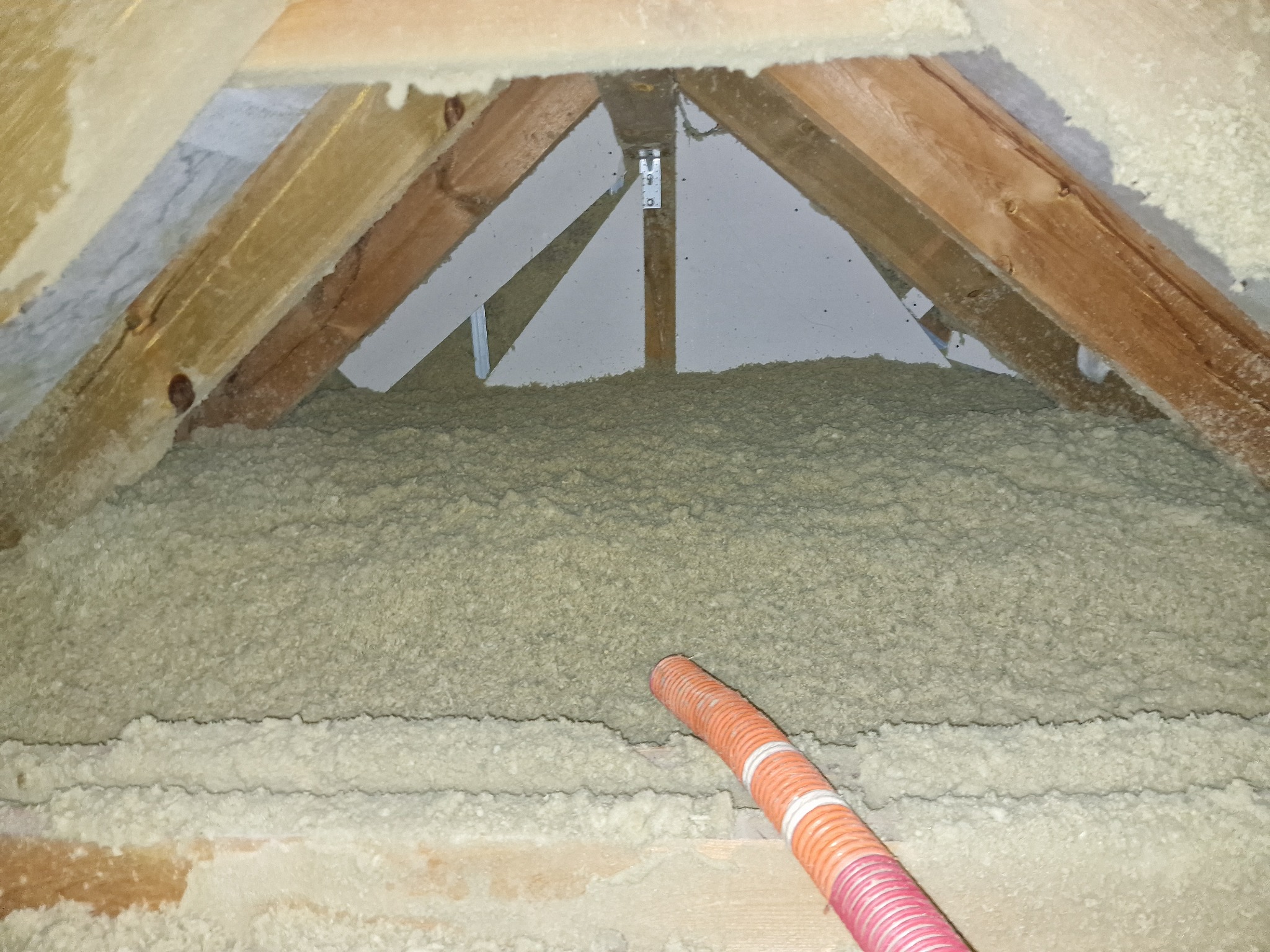 Poddasze z drewnianą konstrukcją krokwi wypełnione szarą, sypką izolacją, z widocznym wężem do jej aplikacji.