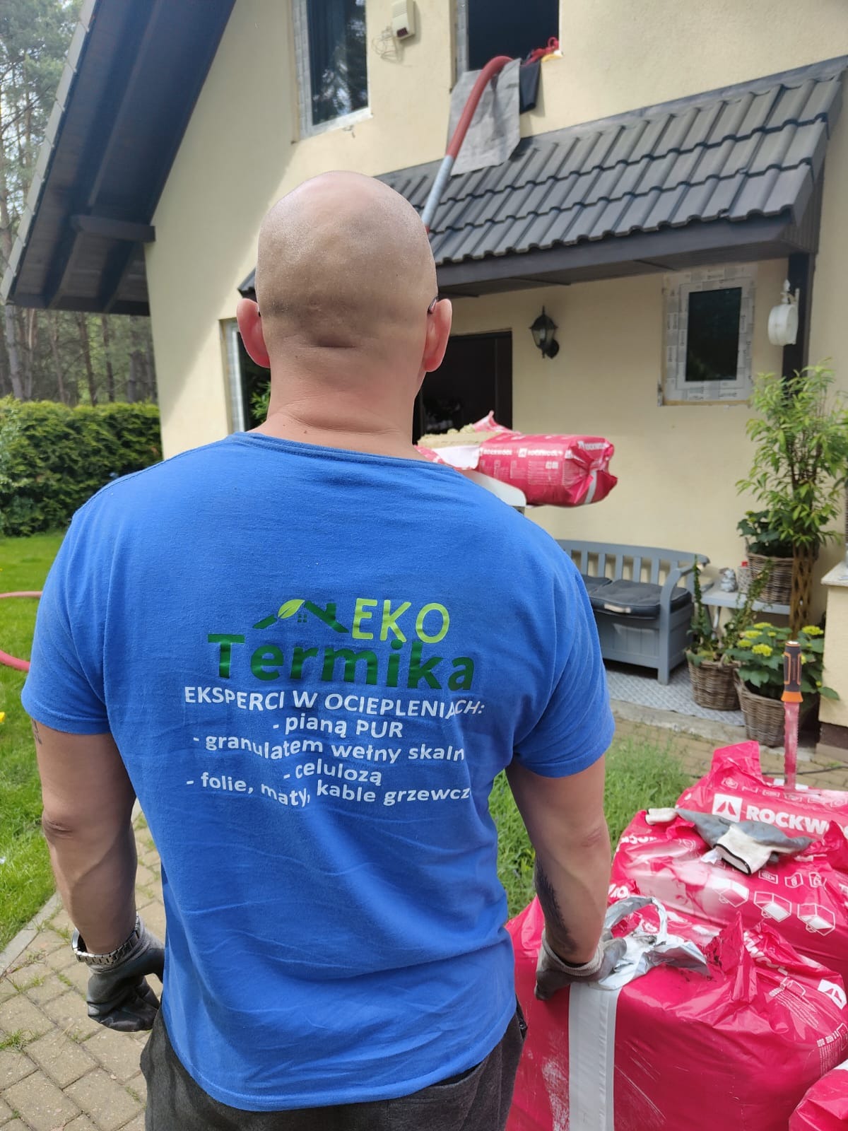Mężczyzna w niebieskiej koszulce z logo firmy ociepleniowej, stojący tyłem do kamery, na tle ocieplanego domu z widocznymi workami materiałów izolacyjnych Rockwool.