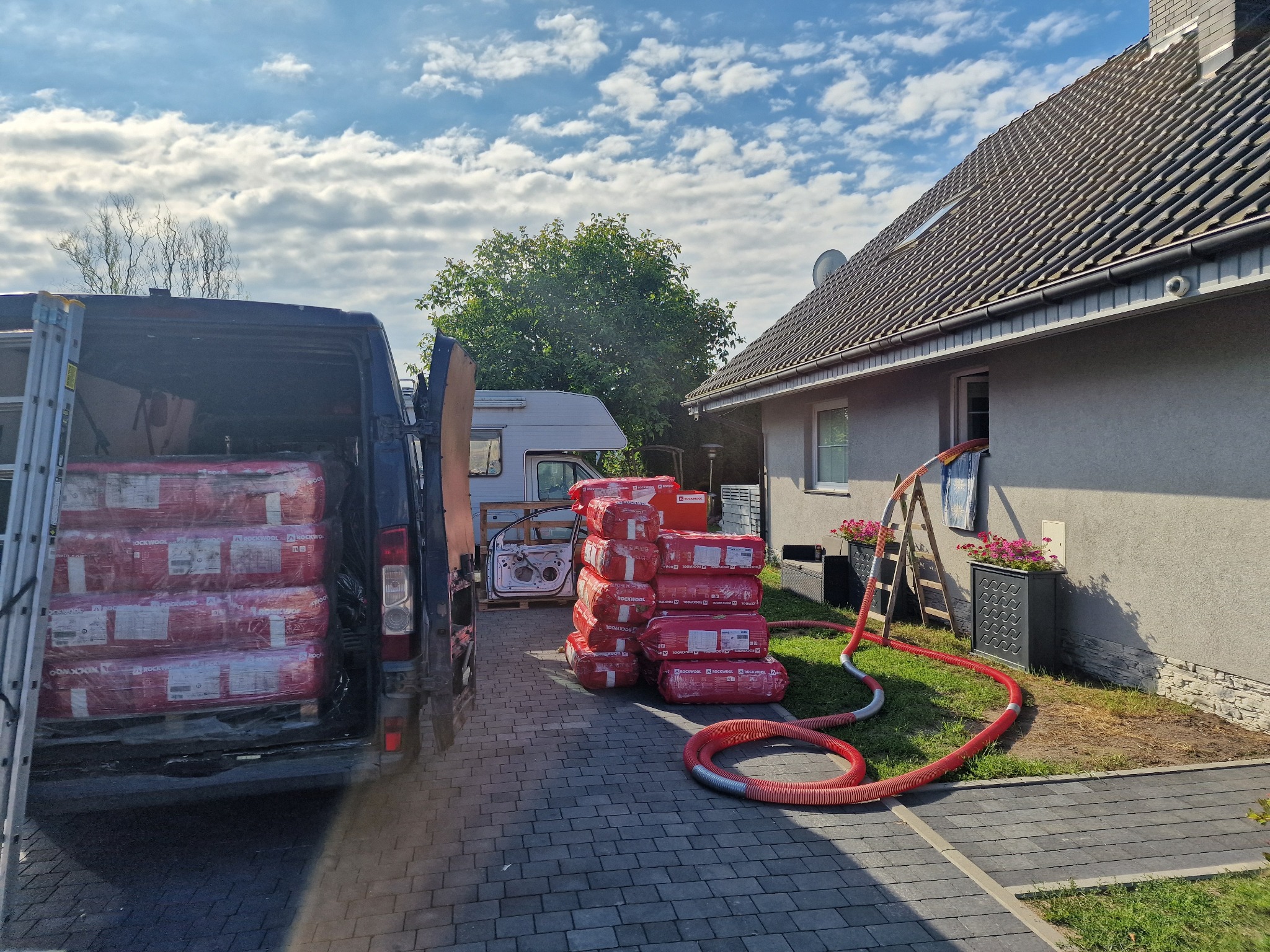Pakiety wełny mineralnej Rockwool w samochodzie dostawczym i na podjeździe przed domem, obok leży czerwony wąż do podawania materiału izolacyjnego, drabina oparta o ścianę budynku.