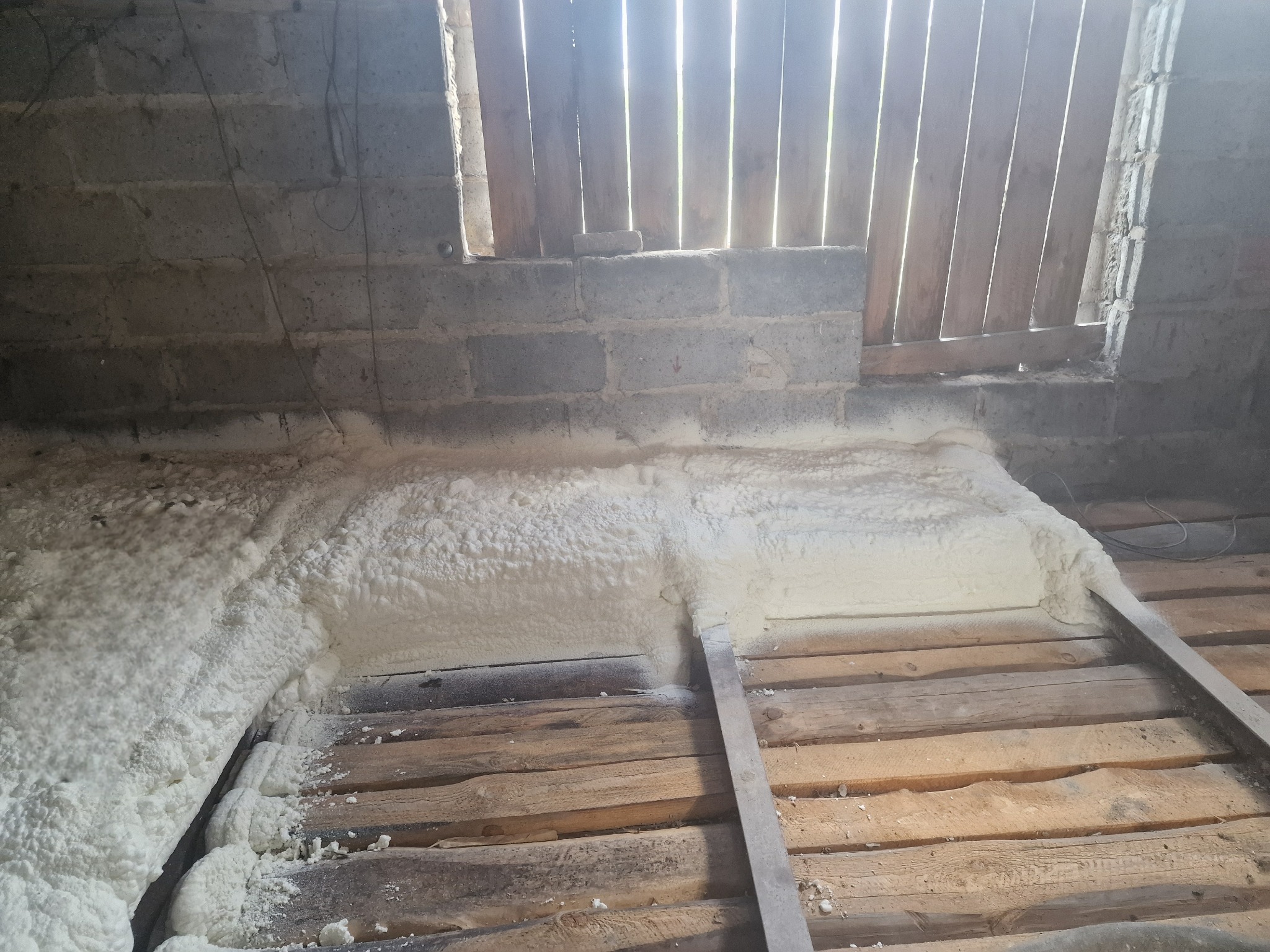 Poddasze z drewnianą konstrukcją i fragmentem ściany z pustaków, częściowo pokryte białą pianką izolacyjną, widoczne szczeliny w deskach dające dostęp światła.