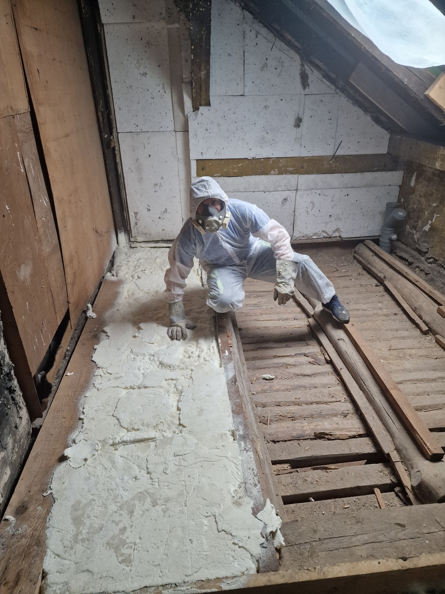 Pracownik w kombinezonie ochronnym i masce aplikuje piankę izolacyjną na poddaszu, widoczne drewniane elementy konstrukcji dachu i podłogi.