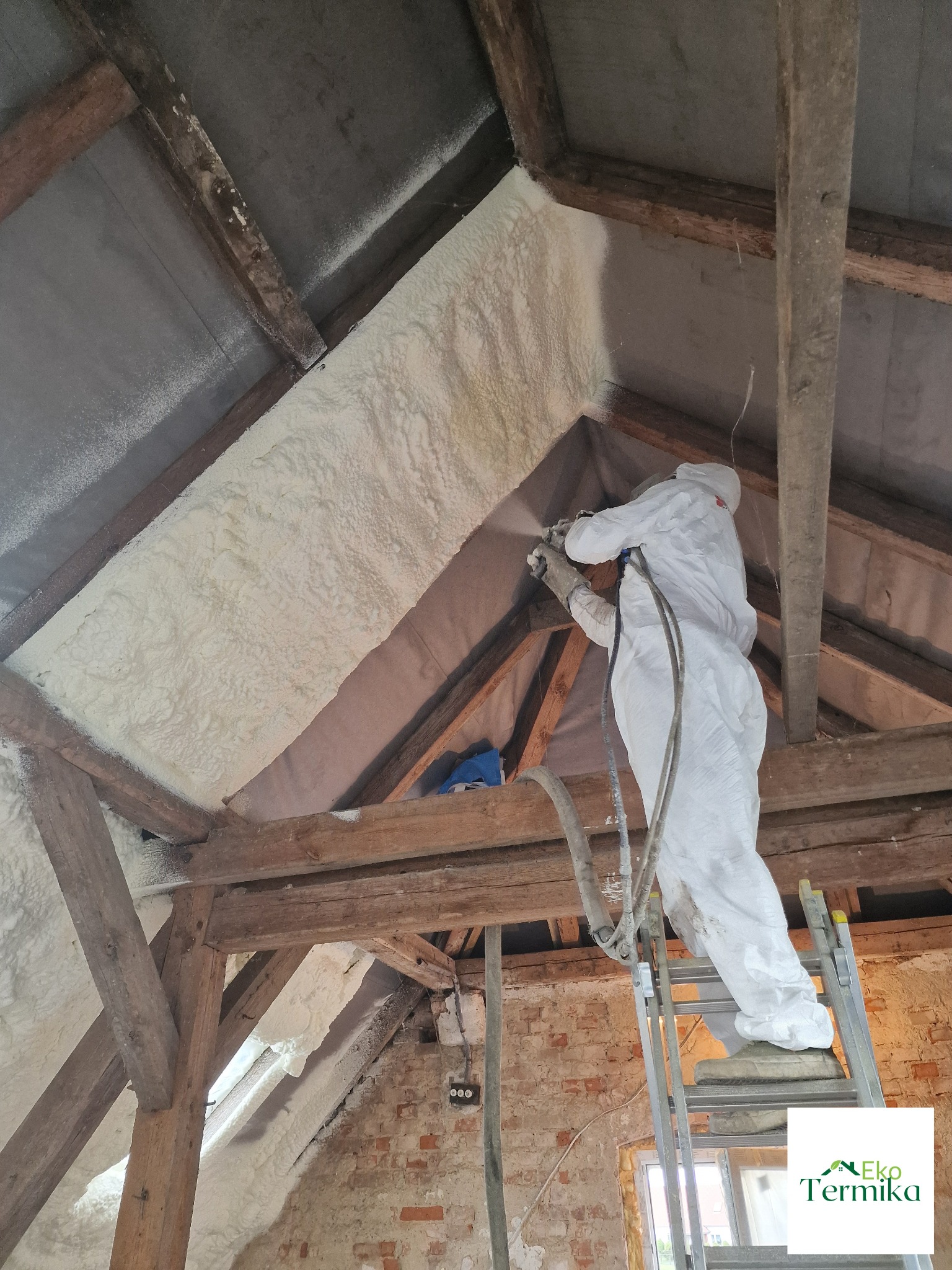 Pracownik w kombinezonie ochronnym aplikuje natryskowo białą pianę izolacyjną na poddaszu z drewnianą konstrukcją, widoczna ściana z cegły i drabina.