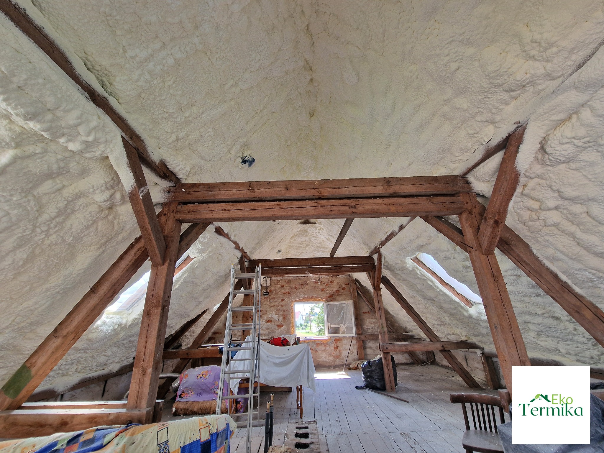 Poddasze z drewnianą konstrukcją pokryte warstwą białej pianki izolacyjnej, widoczne okno dachowe i drabina oparta o ścianę z cegły, w tle logo firmy.