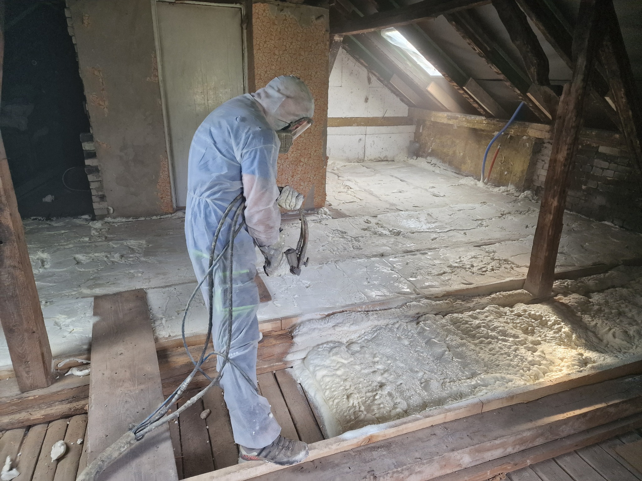 Pracownik w kombinezonie ochronnym aplikuje piankę izolacyjną na poddaszu, widoczne drewniane belki konstrukcyjne i fragmenty starego muru.