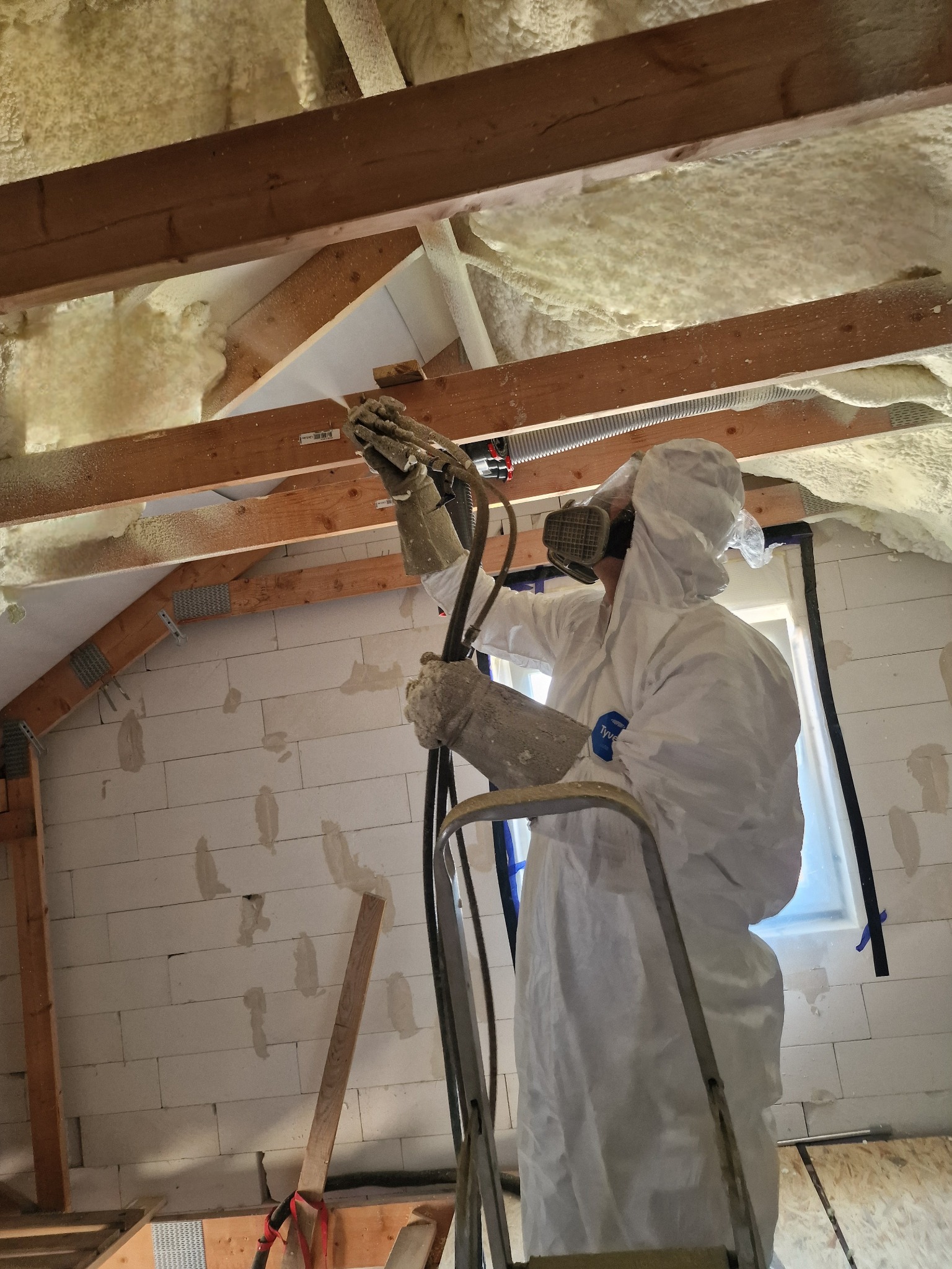 Pracownik w kombinezonie ochronnym i masce na drabinie aplikuje piankę izolacyjną PUR na poddaszu, widoczne drewniane elementy konstrukcyjne i fragment okna.