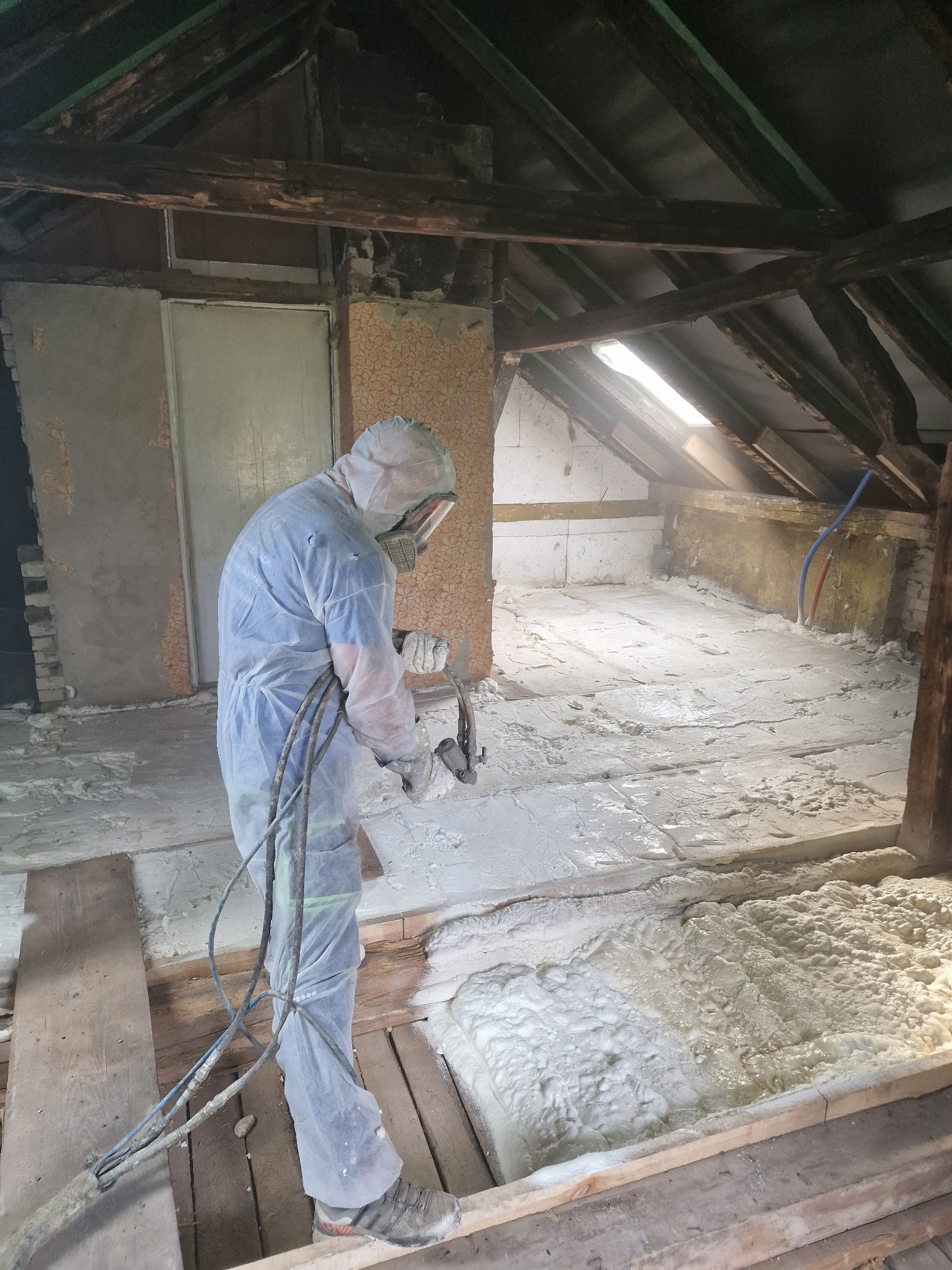 Pracownik w kombinezonie ochronnym aplikuje piankę PUR na poddaszu, widoczne drewniane elementy konstrukcji dachu i fragmenty ścian, jasne światło wpada przez okno dachowe.