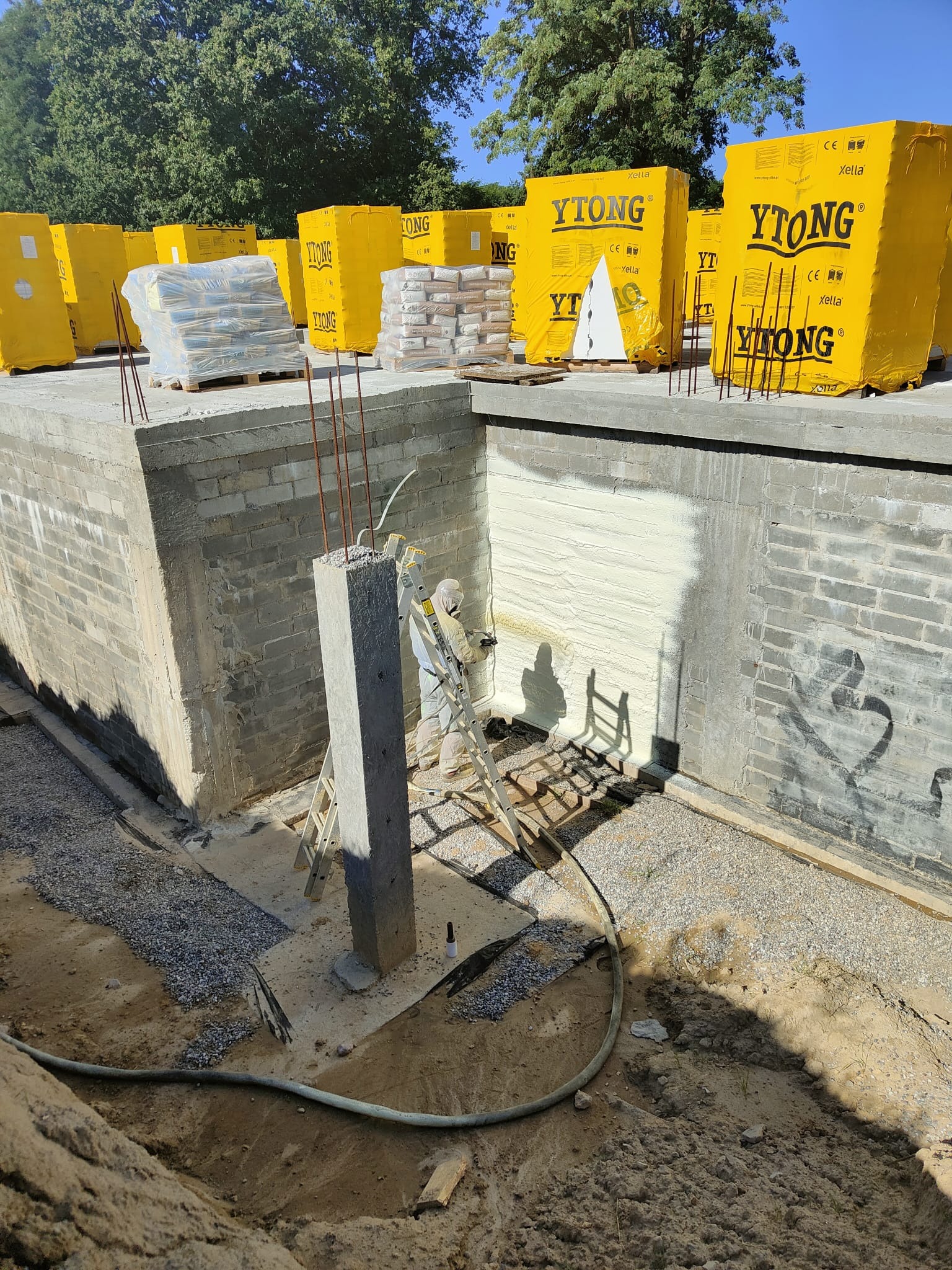 Fundament budynku w trakcie ocieplania pianką natryskową; widoczne żółte opakowania materiałów budowlanych Ytong, pracownik w kombinezonie ochronnym na drabinie, świeżo nałożona warstwa izolacji.