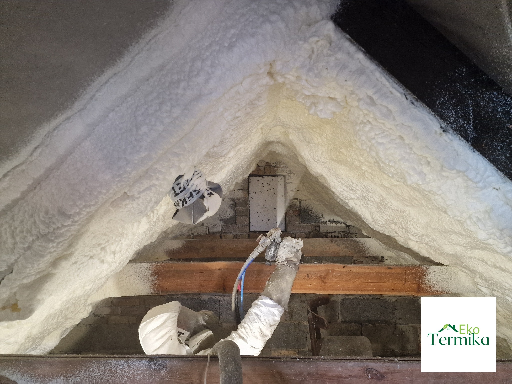 Poddasze z widoczną aplikacją białej pianki izolacyjnej, aplikator w rękawicach ochronnych, fragment logo firmy w prawym dolnym rogu.