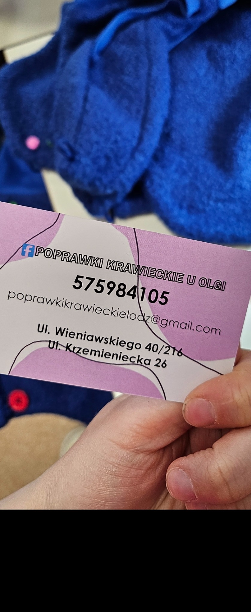 Wizytówka z danymi kontaktowymi do osoby oferującej usługi krawieckie, trzymana w dłoni na tle niebieskiego materiału z ozdobnymi guzikami.