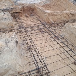 KRIS-bud ROBERT SERVACHAK - Zbrojenie fundamentów w wykopie, widok z góry. Stalowe pręty tworzą siatkę na piasku, przygotowane do zalania betonem. Wykopane ściany z piasku i ziemi.