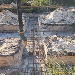 KRIS-bud ROBERT SERVACHAK - Zbrojenie fundamentów pod dom jednorodzinny w Markach. Widoczna rura do wylewania betonu i ułożone cegły. Wykopane rowy i przygotowany teren pod budowę.