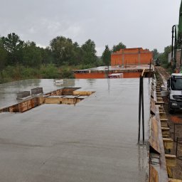 KRIS-bud ROBERT SERVACHAK - Zalany betonem strop budynku w trakcie budowy, widoczne szalunki i wystające pręty zbrojeniowe. W tle budynek z czerwonej cegły i betonowóz.