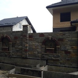 KRIS-bud ROBERT SERVACHAK - Kamienny mur z ceglanymi akcentami i łukami, w tle dwa domy w różnym stanie budowy. Widoczne szalunki fundamentów.