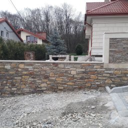 KRIS-bud ROBERT SERVACHAK - Kamienny murek z piaskowca w trakcie budowy, widoczne rusztowania i gruz. W tle dom z czerwoną dachówką i iglaste drzewo. Ujęcie z poziomu gruntu.