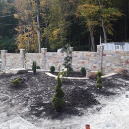 KRIS-bud ROBERT SERVACHAK - Kamienny mur z kolumnami i nowo posadzone rośliny ozdobne na tle lasu. Widoczny fragment kamiennego chodnika i paleta z płytami.
