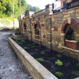 KRIS-bud ROBERT SERVACHAK - Kamienny mur z dekoracyjnymi wnękami i rzeźbami zwierząt, oddzielający posesję od drogi. Wzdłuż muru rabata z nasadzeniami. Widoczne elementy wykończenia z cegły.