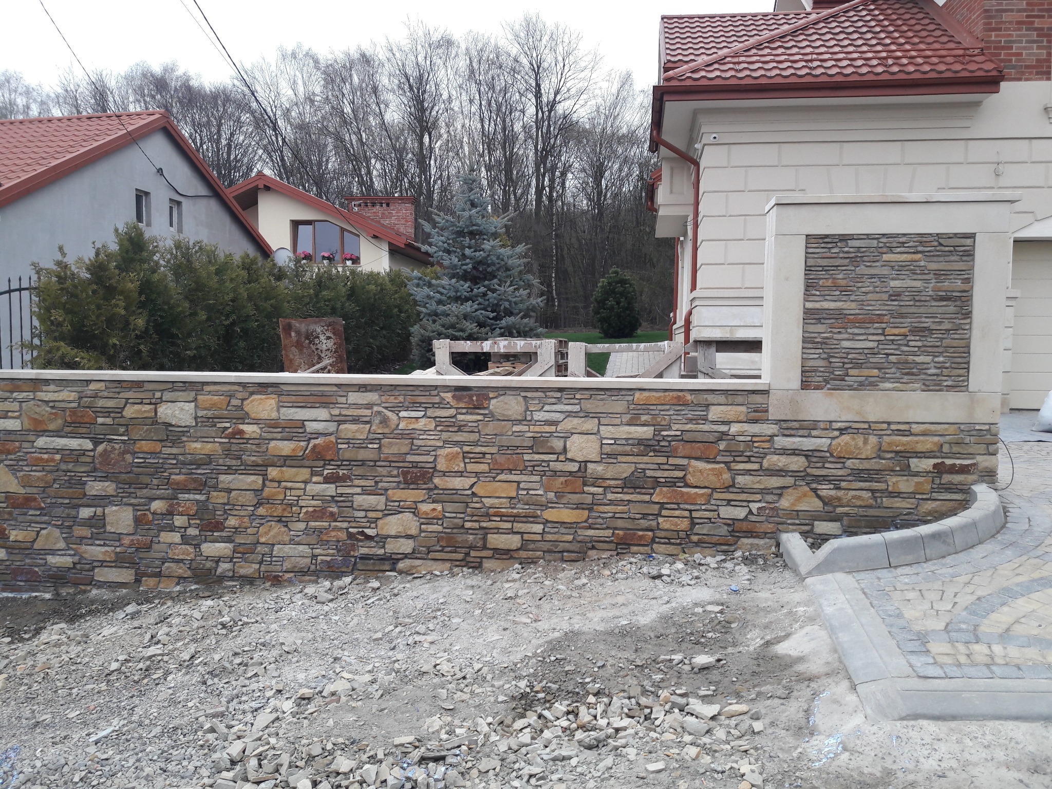 Kamienny murek z piaskowca w trakcie budowy, widoczne rusztowania i gruz. W tle dom z czerwoną dachówką i iglaste drzewo. Ujęcie z poziomu gruntu.