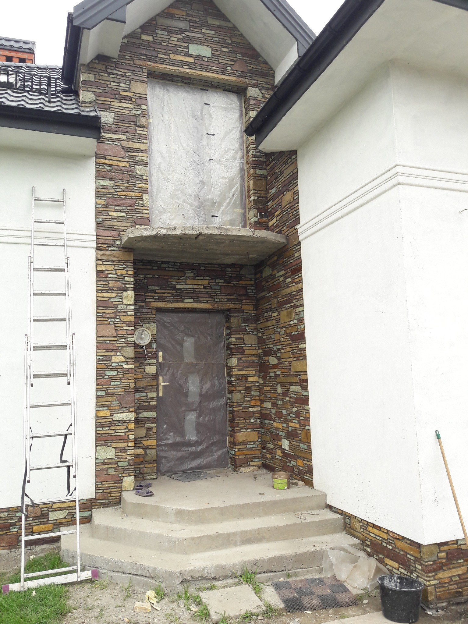 Fragment elewacji domu z kamienia naturalnego w trakcie prac wykończeniowych. Widoczna drabina, zabezpieczone okno i drzwi folią. Część ściany otynkowana na biało.