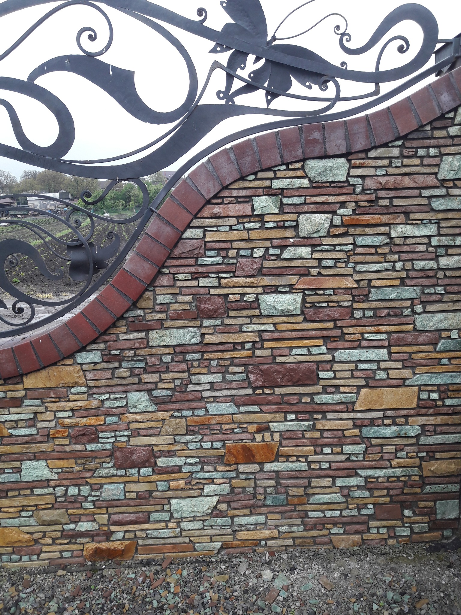 Kamienny mur z dekoracyjnym, kutym elementem bramy. Różnokolorowe kamienie tworzą mozaikę, a ceglana obwódka dodaje charakteru. Kuta brama z motywem roślinnym.