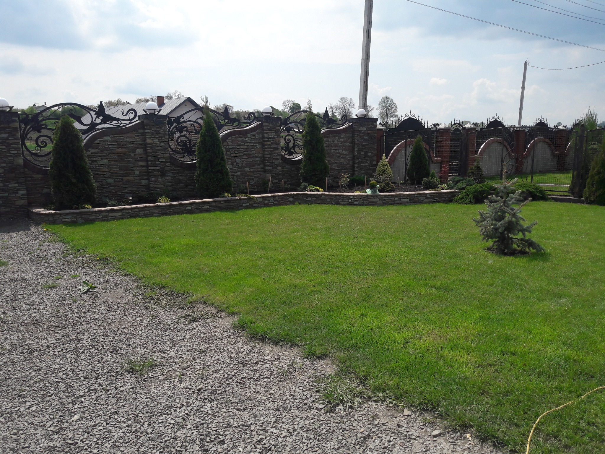 Kamienny mur z kutymi elementami i tujami oddziela trawnik od żwirowej ścieżki. W tle widoczny dom i słup energetyczny.