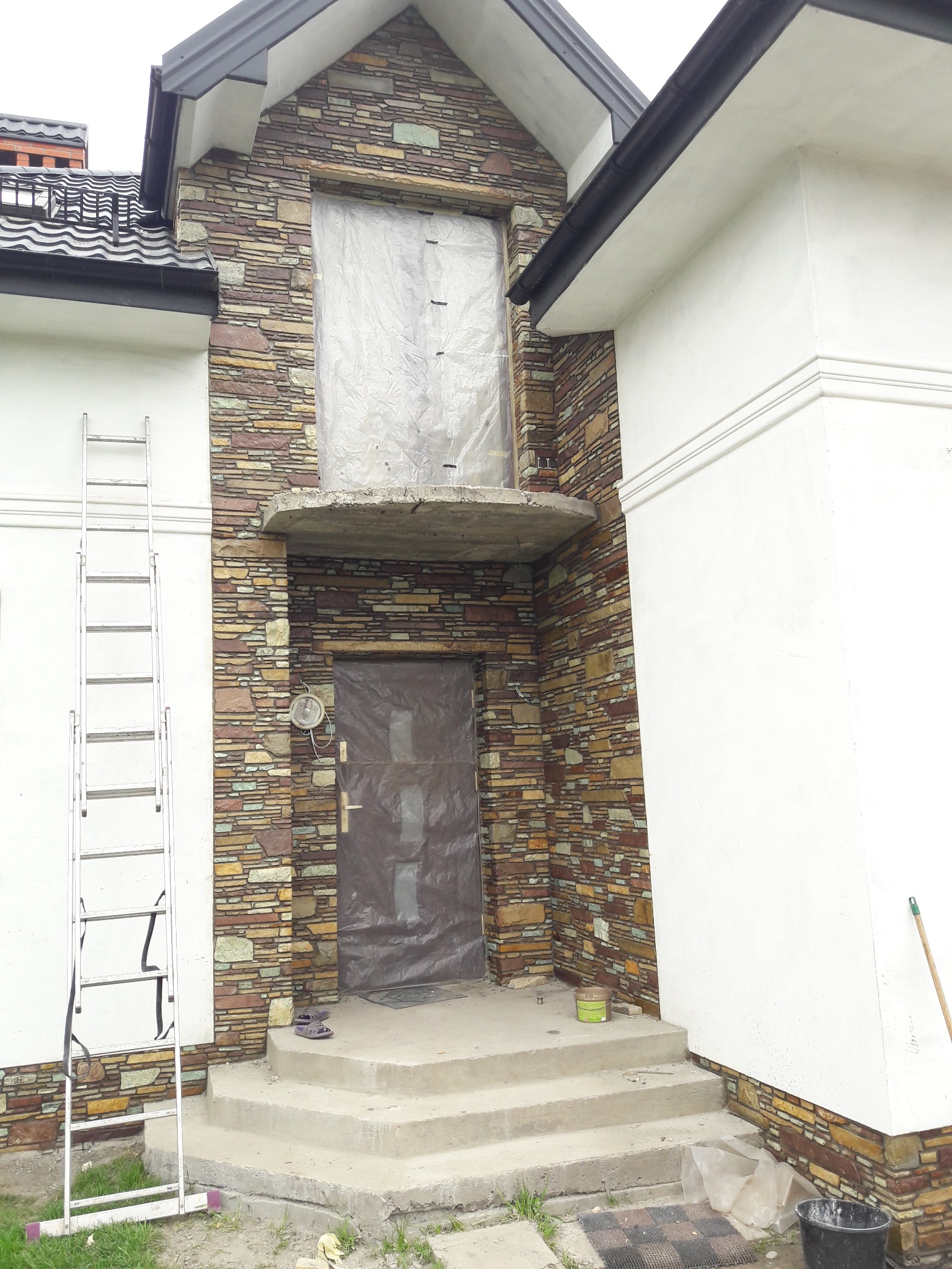 Fragment domu z elewacją z kamienia dekoracyjnego, częściowo zakrytą folią ochronną. Widoczna drabina oparta o ścianę i schody wejściowe. Detal wykończenia elewacji.